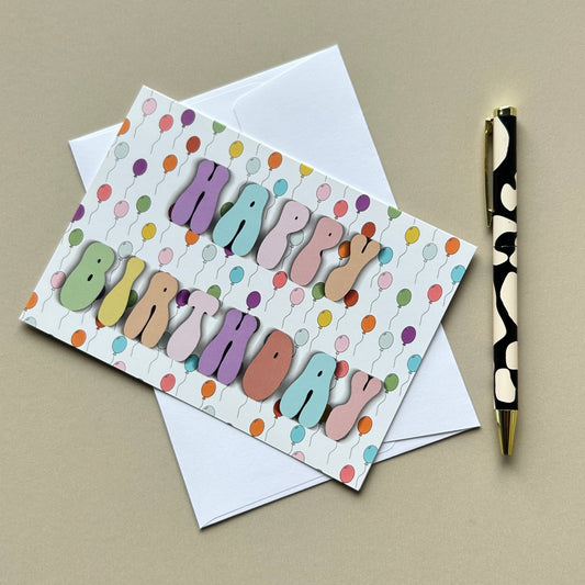 A colorful 'Happy Birthday' greeting card with pastel balloon-style letters and a white envelope, styled on a beige surface next to a black-and-gold pen.