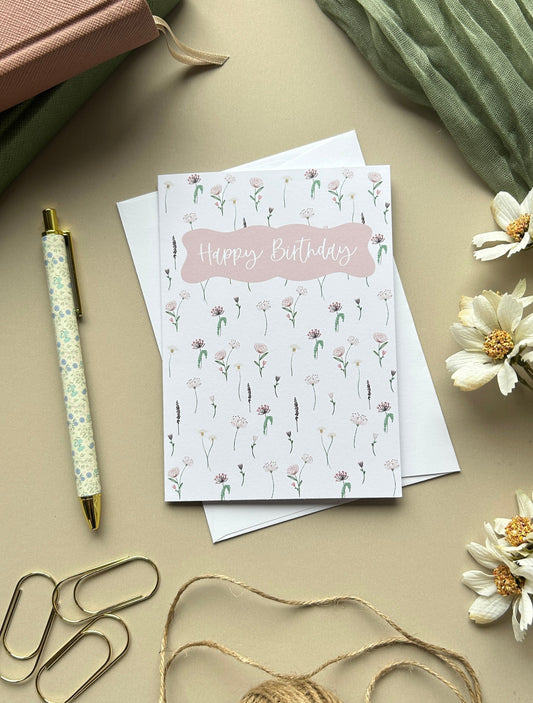 A styled flat-lay featuring a 'Happy Birthday' greeting card with a pink floral pattern, surrounded by a pen, paperclips, twine, flowers, and notebooks on a beige background.