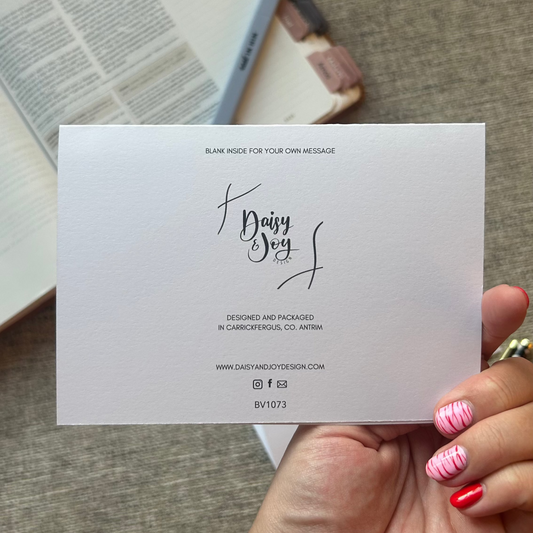 A hand holding a blank white card with elegant black script text, set against a cozy background of an open book and a textured gray fabric surface.