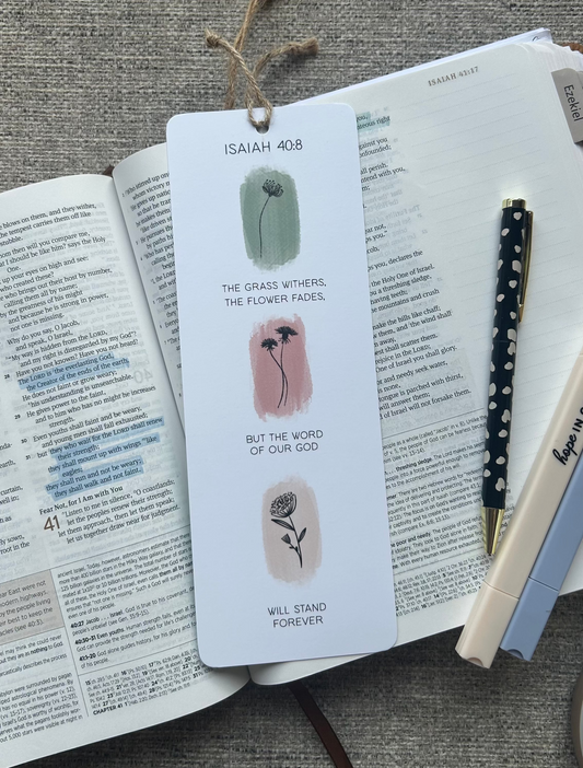 A white paper bookmark with illustrations and text from Isaiah 40:8 lies on an open book, styled with a pen and another pen on a textured gray fabric background.