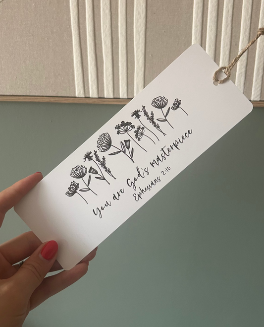 A hand with red nail polish holds a white rectangular bookmark or tag printed with black floral line art and the text 'You are God's masterpiece John 3:30'. The background shows a styled interior wall with cream curved molding and a framed photo.