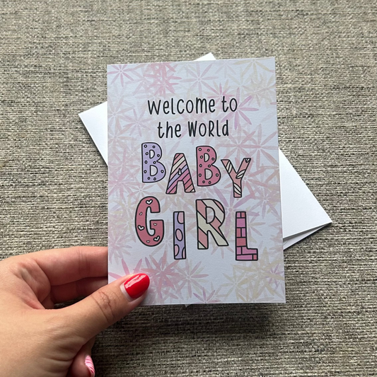 A hand with pink nail polish holds a square baby shower card that reads 'Welcome to the World BABY GIRL' in colorful patterned letters, with a white envelope behind it on a textured gray fabric background.