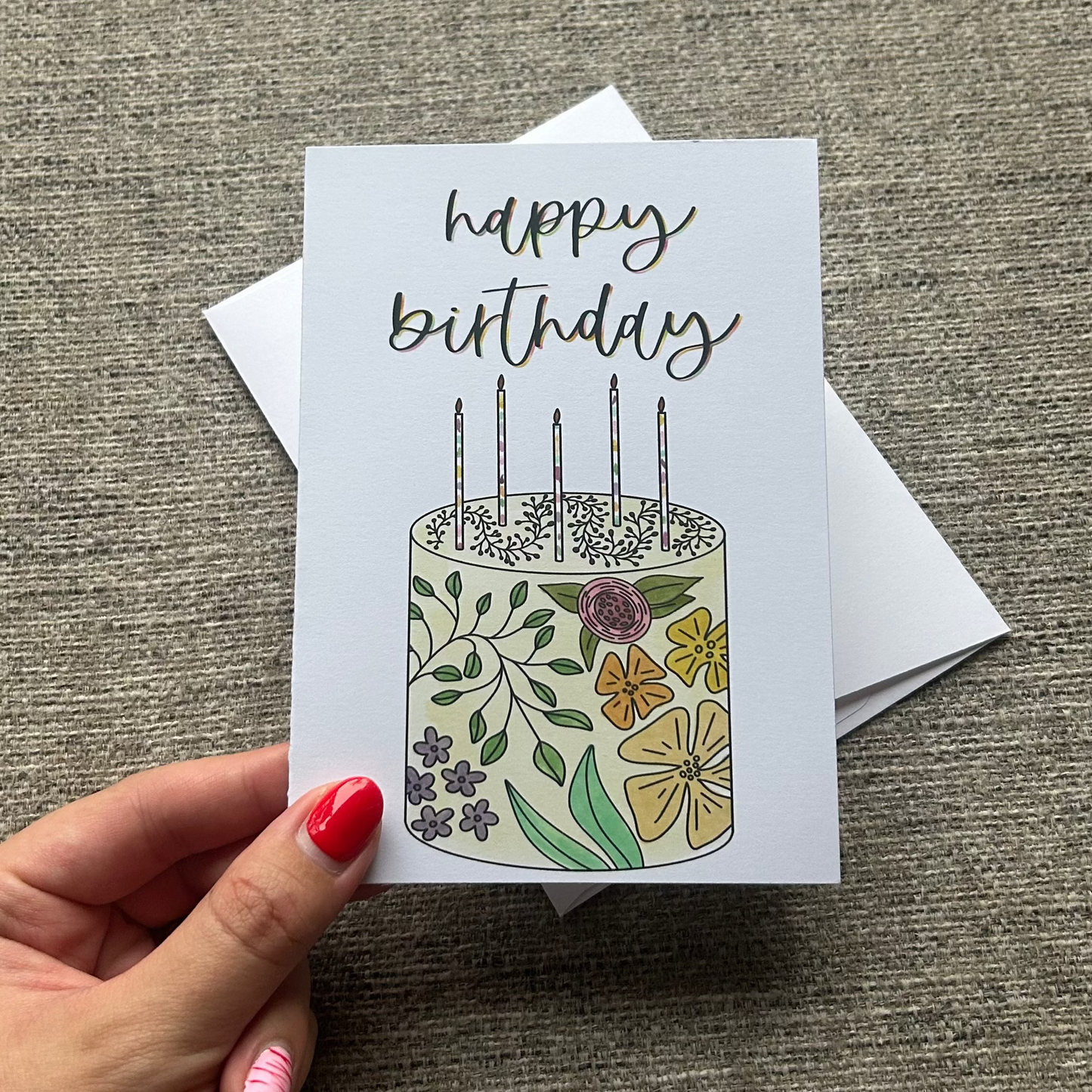 A hand with red nail polish holds a white 'happy birthday' greeting card featuring a floral cake illustration, with white envelopes behind it on a textured gray fabric background.