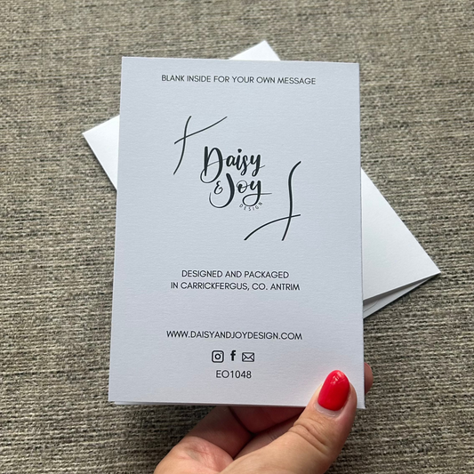 A hand with red nail polish holds a blank white business card template featuring the text 'Dairy Joy' in black script, set against a textured gray fabric background.