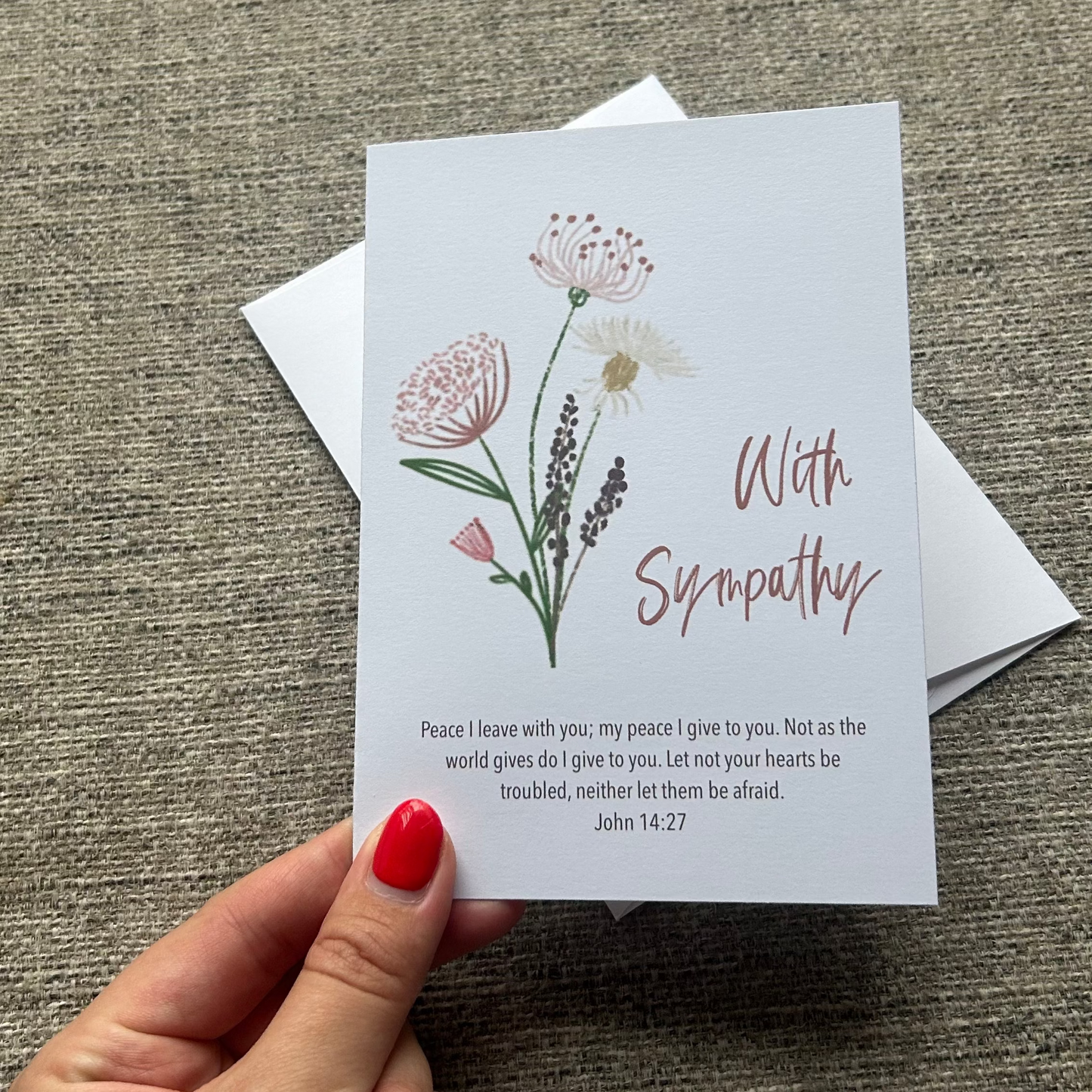 A hand with red nail polish holds a sympathy greeting card featuring delicate pink and white wildflower illustrations and the words 'With Sympathy' on a textured gray fabric background.