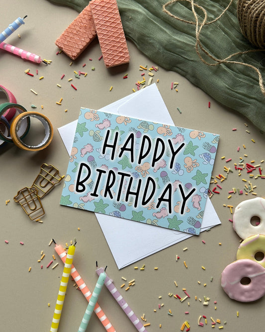 A square birthday card with a pastel under the sea pattern and the words 'HAPPY BIRTHDAY' in black lettering, placed on top of a white envelope. The card is styled in a flat lay with pastel pencils, ribbon, confetti, cookies, and fabric on a neutral surface.
