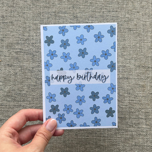 A hand holding a square handmade greeting card with a blue floral pattern and 'happy birthday' written across the center, set against a textured gray fabric background.