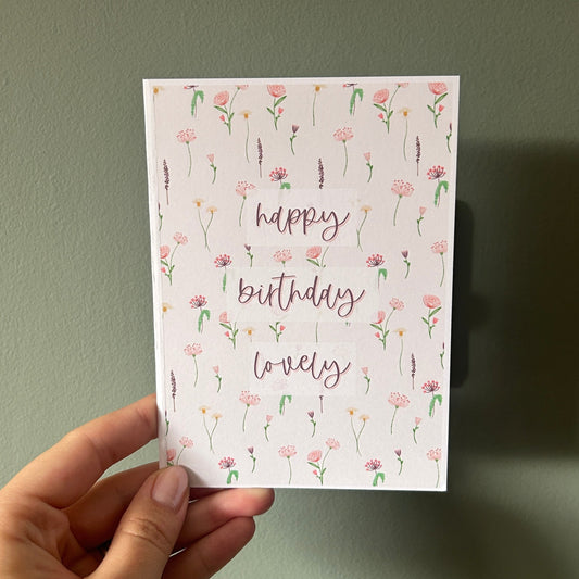 A hand holding a square greeting card with a white background, delicate pink and green floral border, and the words 'happy birthday lovely' in a cursive rose-gold font, set against a muted green wall.