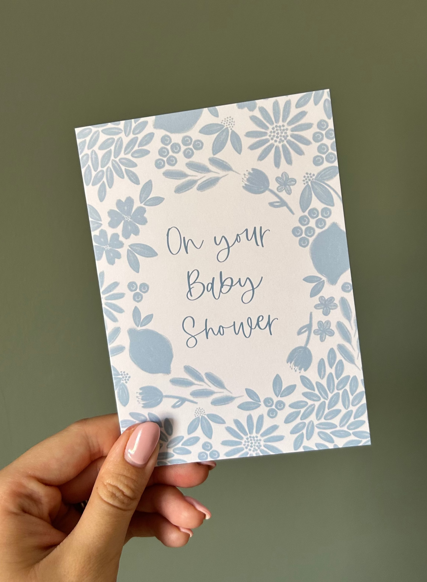 Hand holding a baby shower card with floral design and text on a plain background