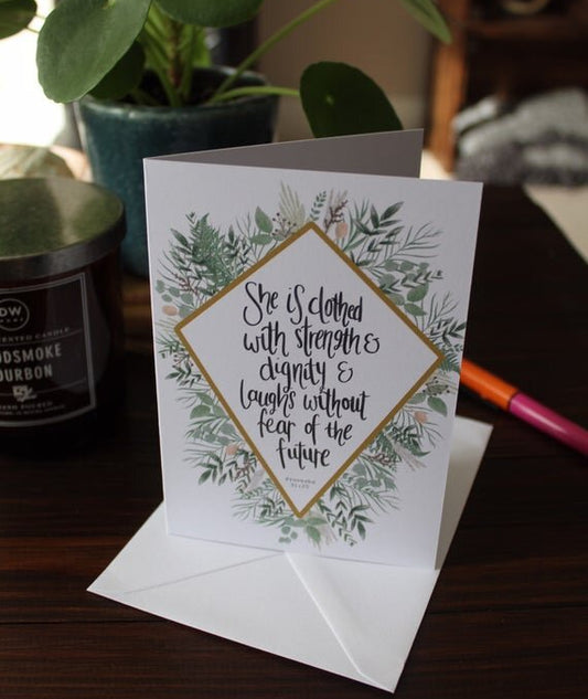 A greeting card with a floral diamond illustration and a bible verse, placed on a wooden surface with a plant and a candle in the background.