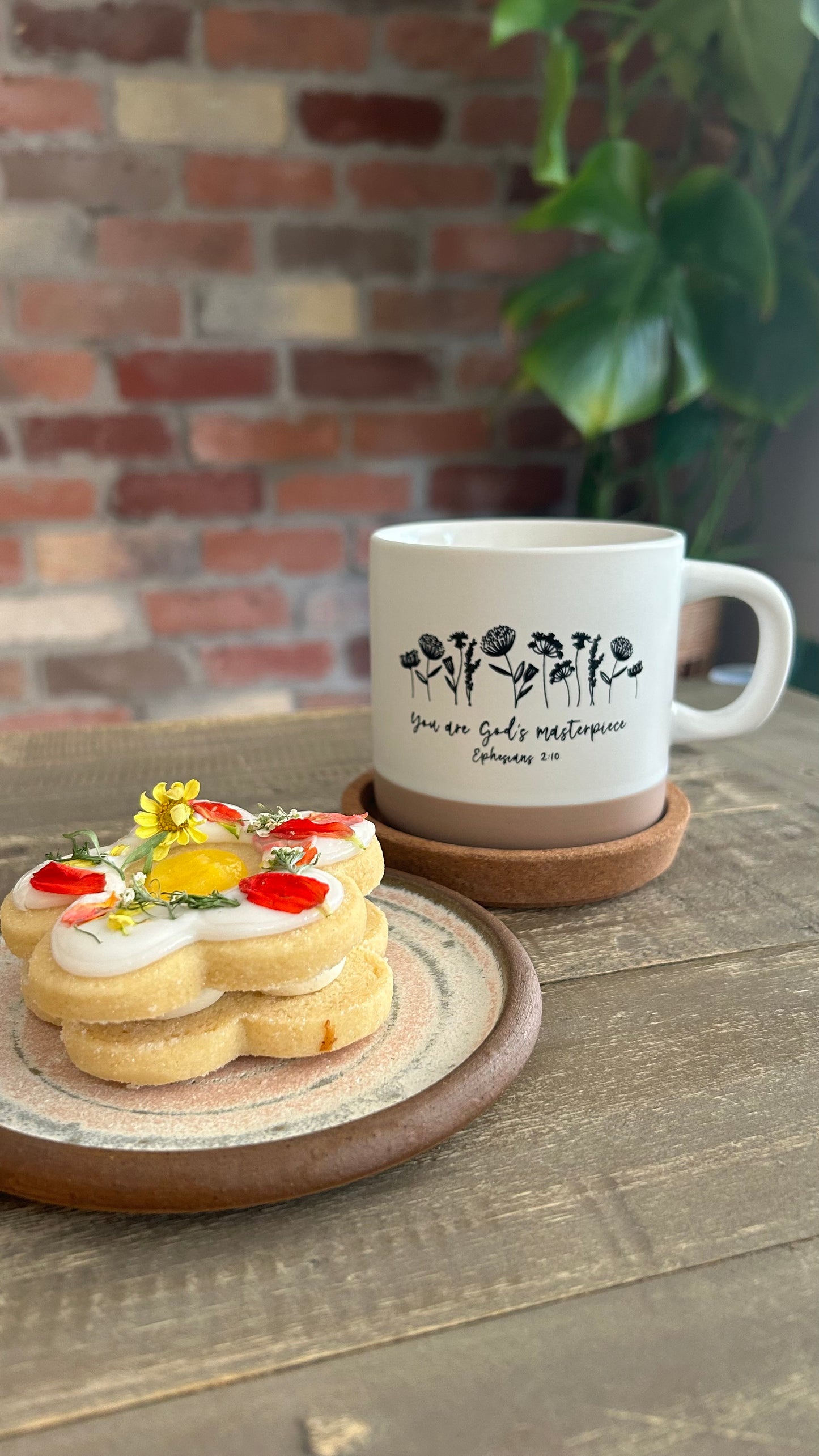 Mug with floral design and text of Ephesians 2:10 on a wooden surface with cookies and a plant in the background