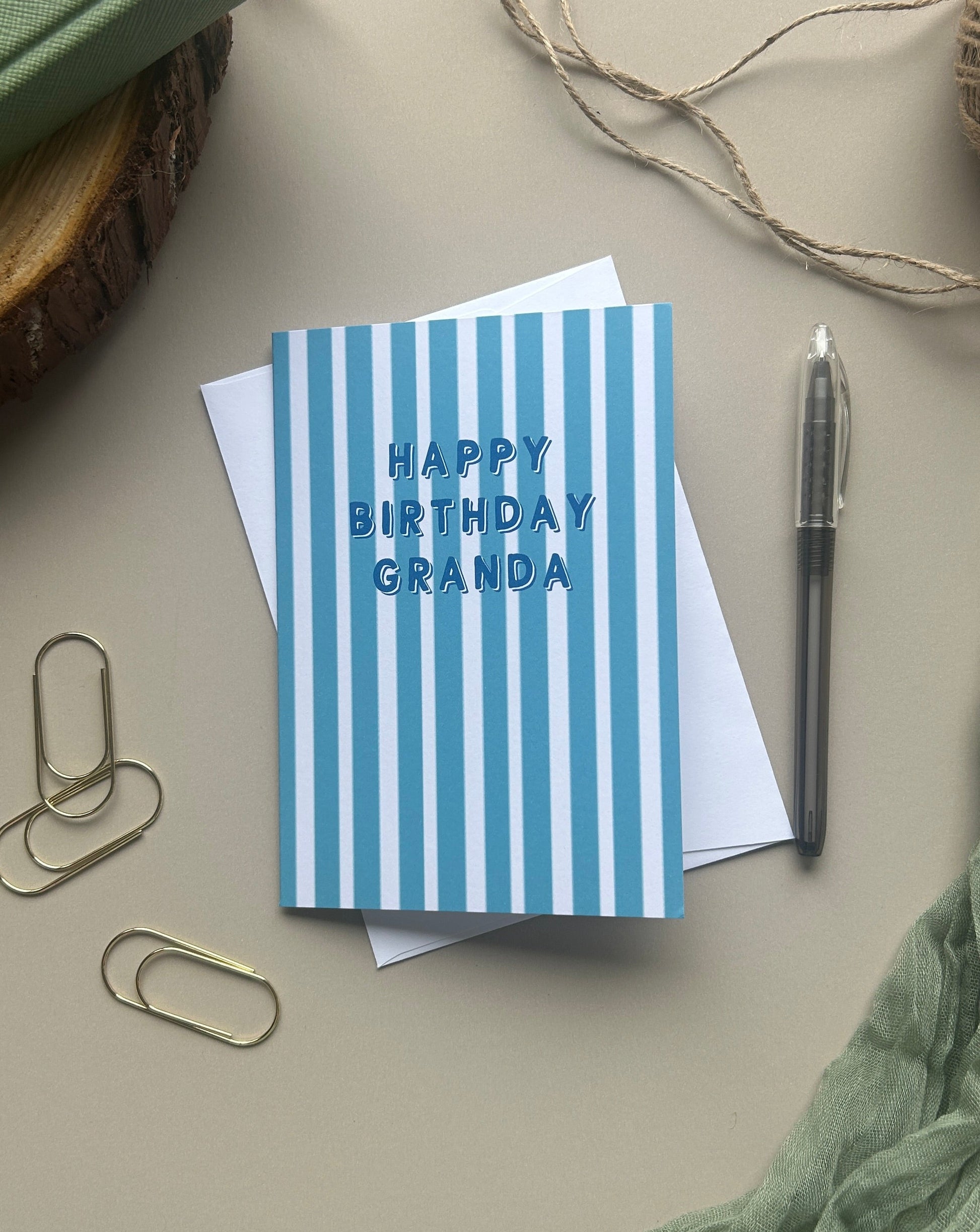 A blue and white striped greeting card that reads 'HAPPY BIRTHDAY GRANDA' in blue lettering, styled in a flat-lay arrangement with a white envelope, a pen, paperclips, twine, and a green notebook on a neutral beige background.