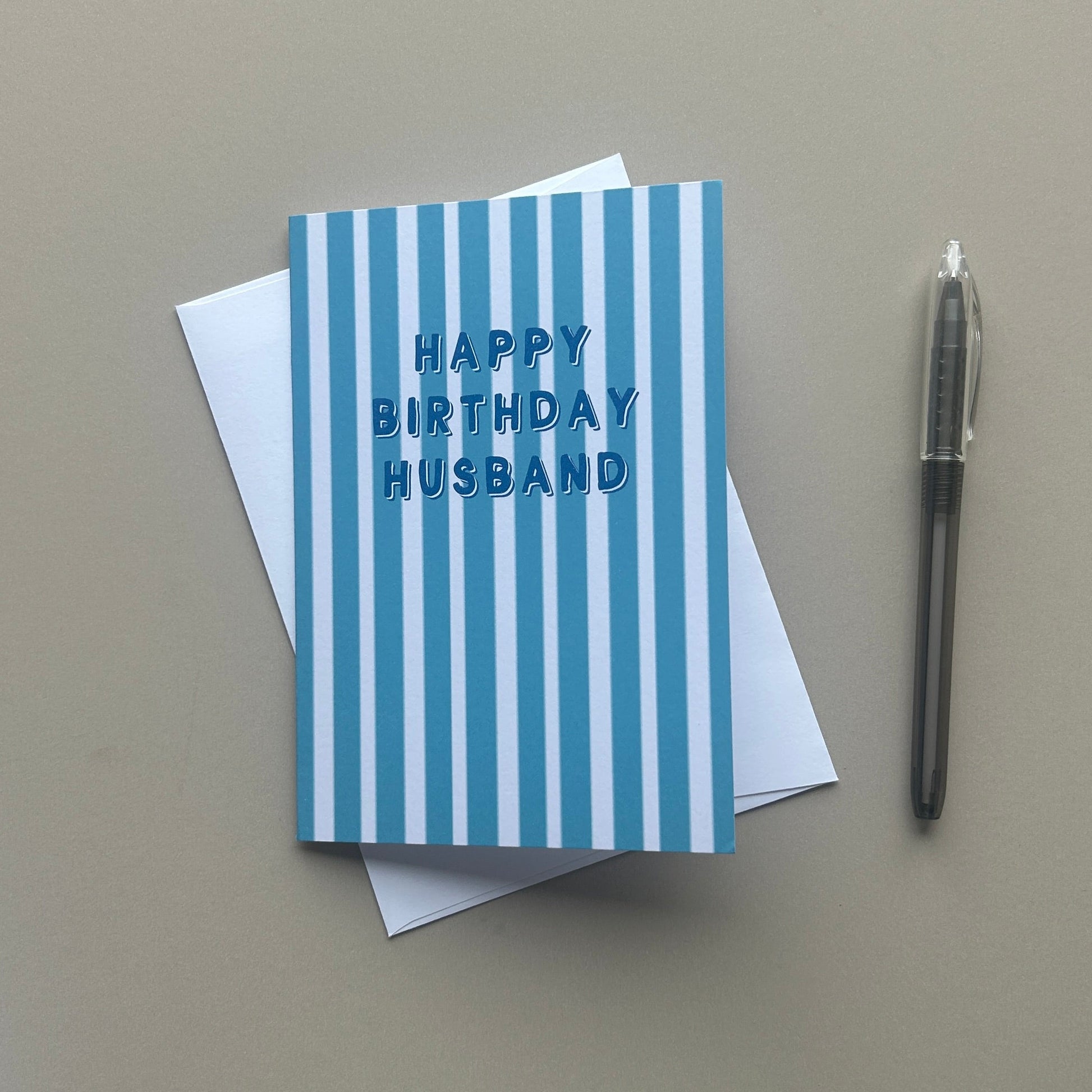 A blue and white striped greeting card with 'HAPPY BIRTHDAY HUSBAND' written in blue, resting on top of a white envelope. A black pen is placed to the right on a solid beige background.