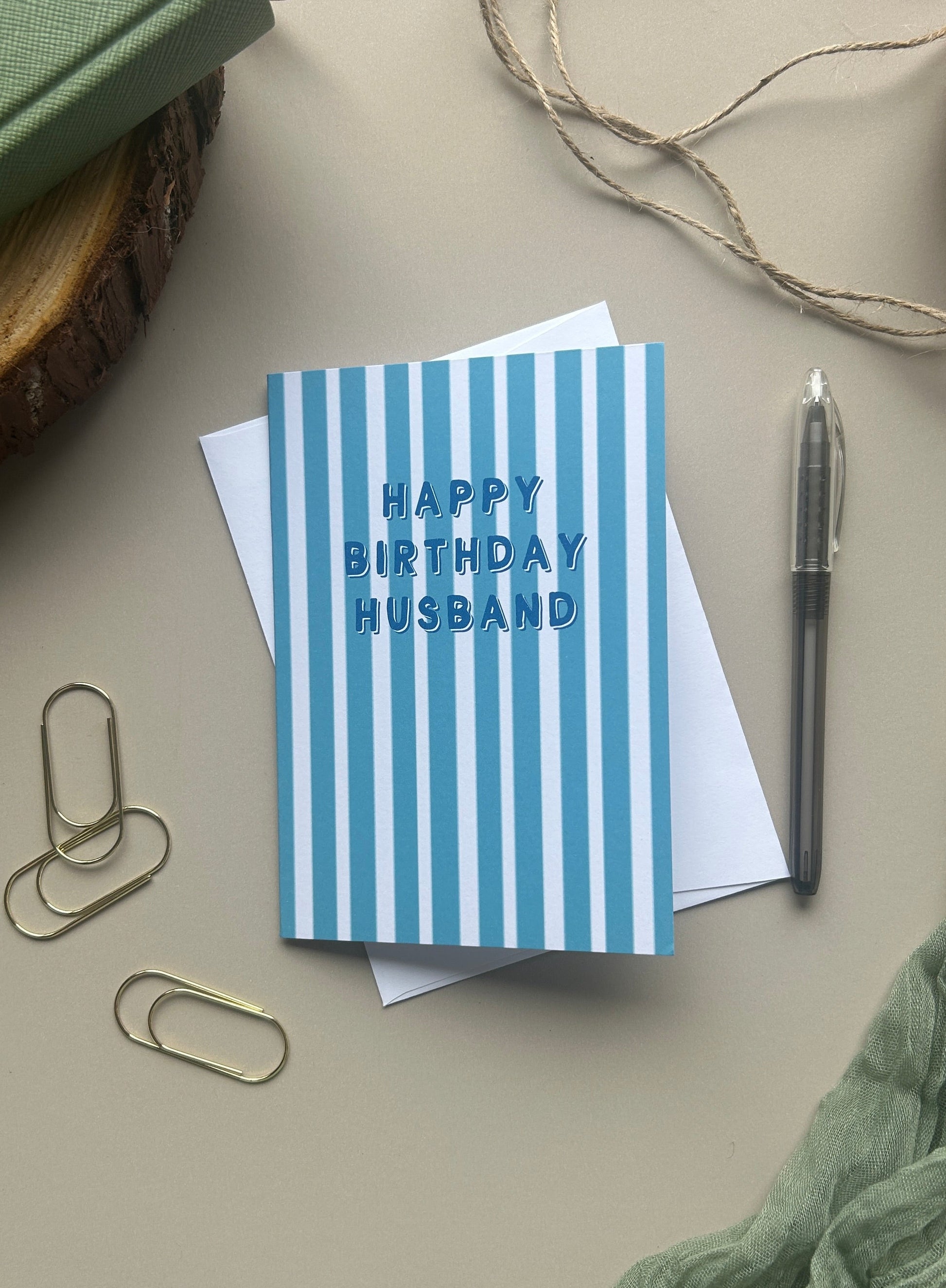 A blue and white striped greeting card with the text 'HAPPY BIRTHDAY HUSBAND' in blue, styled in a flat lay with a white envelope, paperclips, a pen, twine, and a green notebook on a beige background.