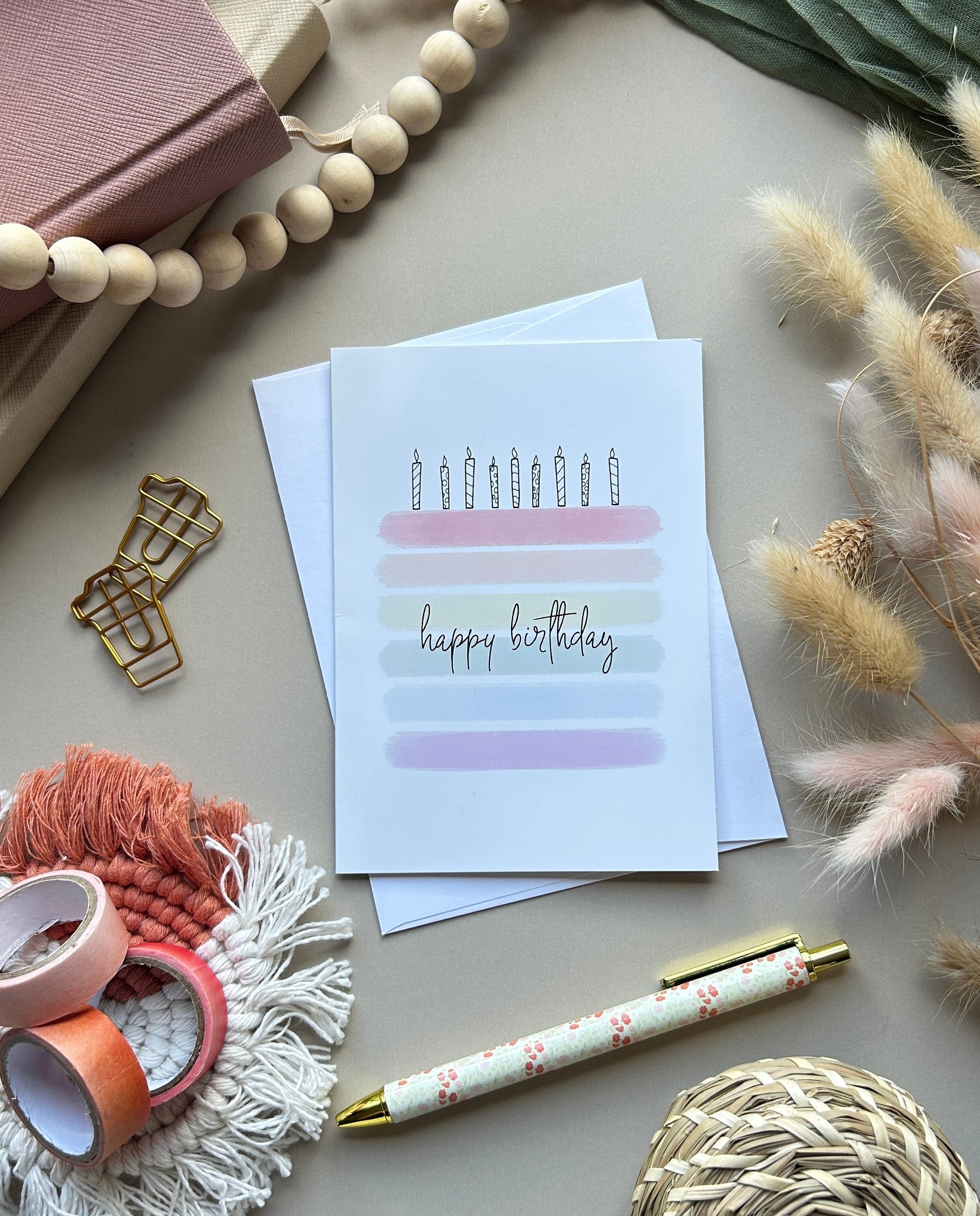 A flat-lay of a birthday greeting card with a simple candle and pastel stripe design reading 'happy birthday'. The card is styled with a pen, washi tape, beads, a woven ball, and dried grass on a neutral background.