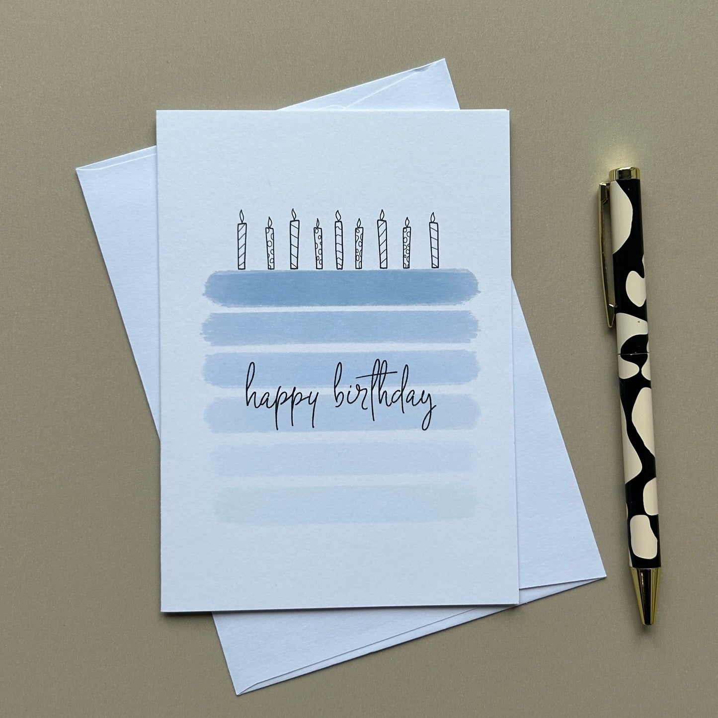 A white birthday greeting card with a simple illustration of candles on a blue-striped birthday cake and the words 'happy birthday' in script, shown with white envelopes and a black-and-white patterned pen on a warm gray background.