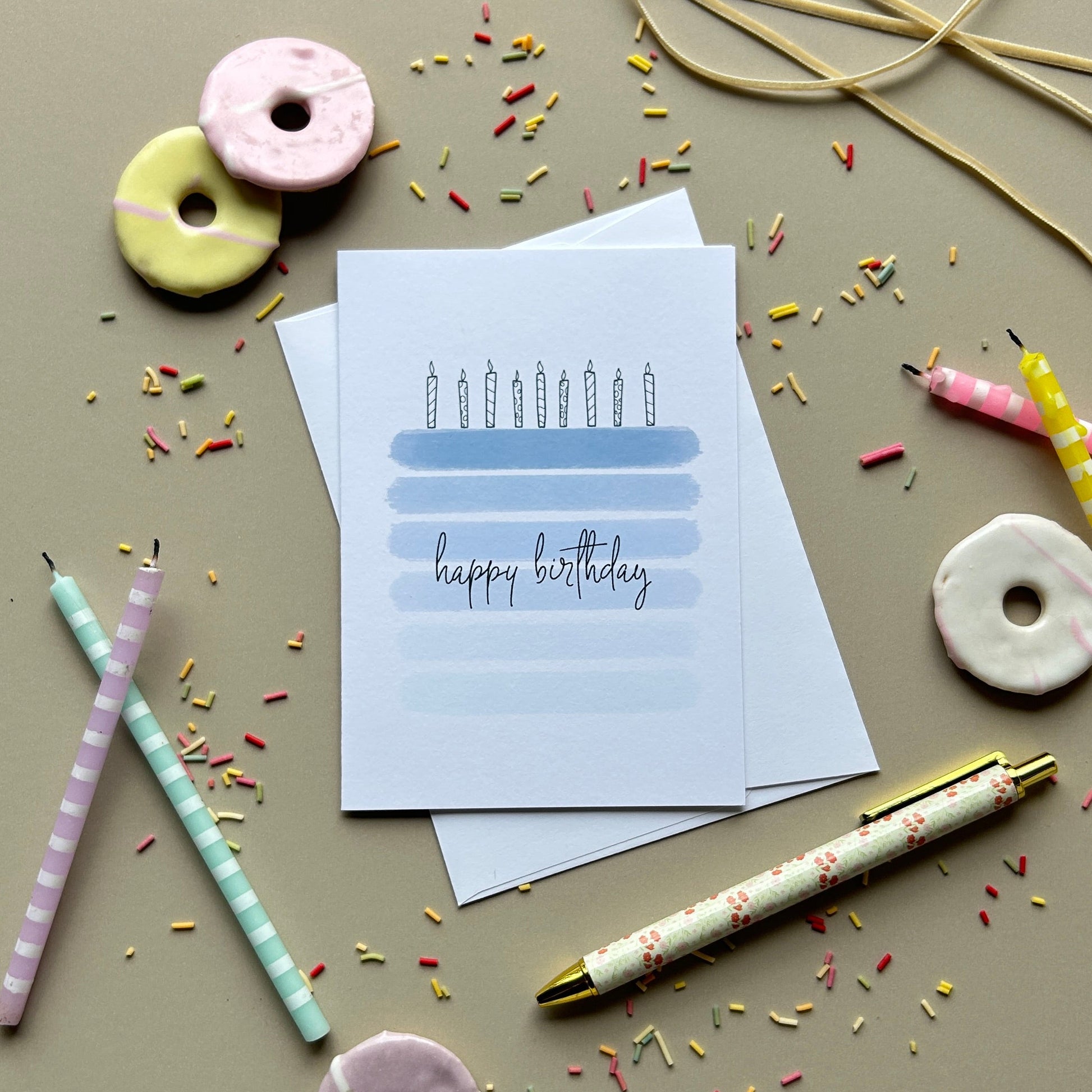 A styled flat-lay of a 'happy birthday' greeting card with a candle illustration, surrounded by pastel donut-shaped candles, striped party straws, and decorative sprinkles on a neutral beige background.