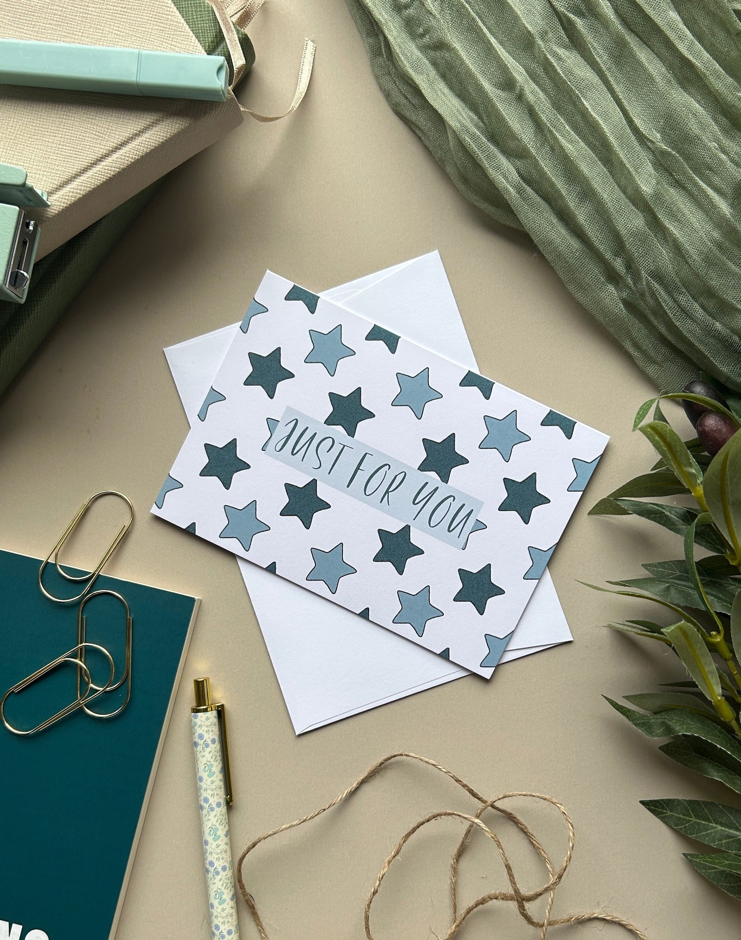 A styled flat-lay featuring a white card with a blue star pattern and the text 'JUST FOR YOU' in script, placed on a beige surface with notebooks, paper clips, twine, and greenery.
