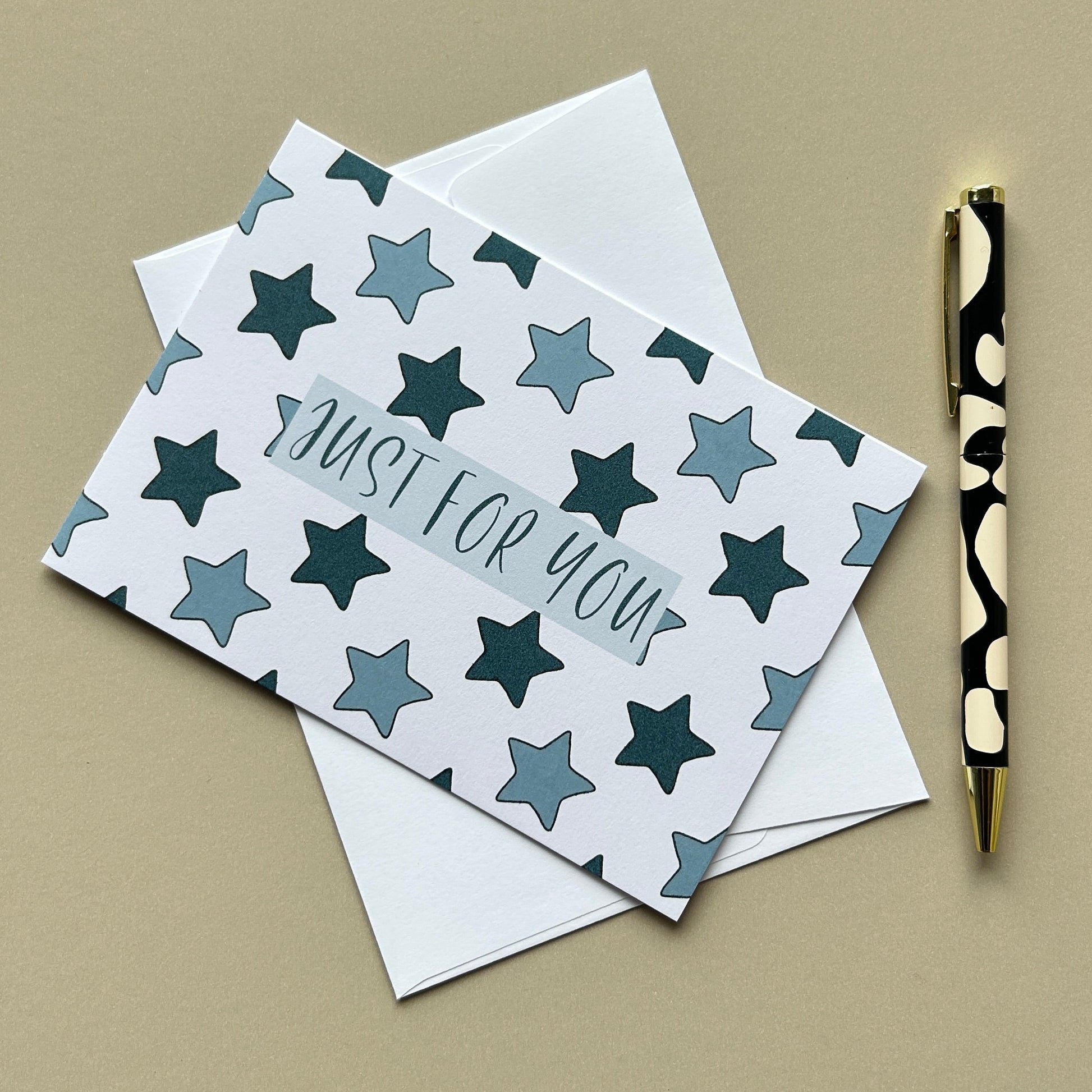 A square greeting card with a white background and a pattern of dark teal and gray stars, featuring the text 'JUST FOR YOU' in a cursive font. The card is shown with a white envelope behind it on a beige background, next to a black and gold patterned pen.