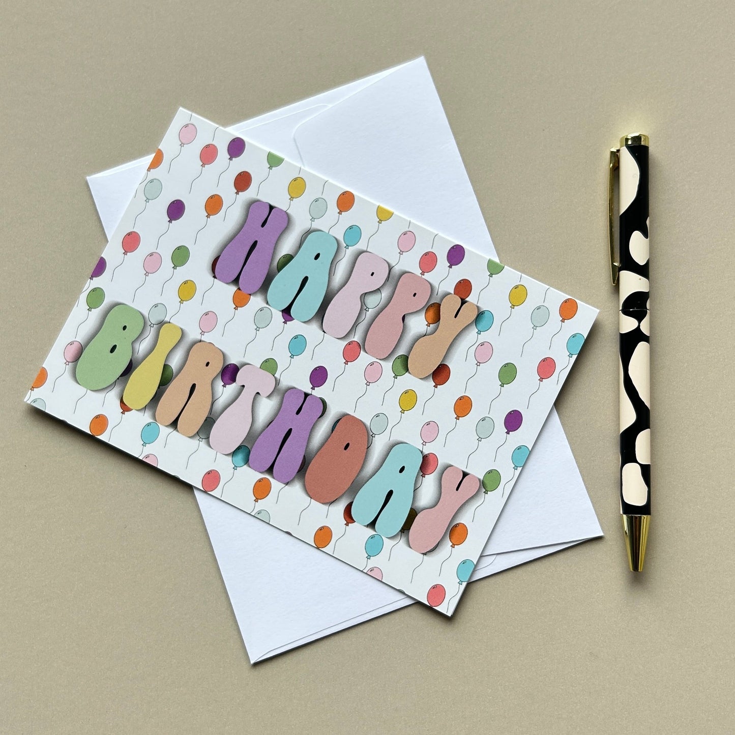 A colorful 'Happy Birthday' greeting card with pastel balloon-style letters and a white envelope, styled on a beige surface next to a black-and-gold pen.