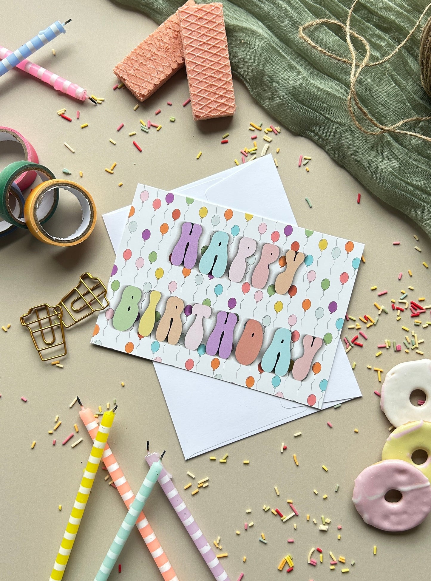 A colorful 'Happy Birthday' greeting card with a white envelope, styled in a flat-lay arrangement with pastel pencils, washi tape, waffle pieces, and donut-shaped candies on a beige surface.