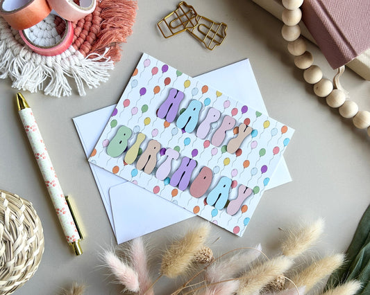 A colorful 'Happy Birthday' greeting card with pastel lettering and confetti, shown with a white envelope on a neutral tabletop surrounded by decorative props including a pen, beads, dried grass, and a woven coaster.