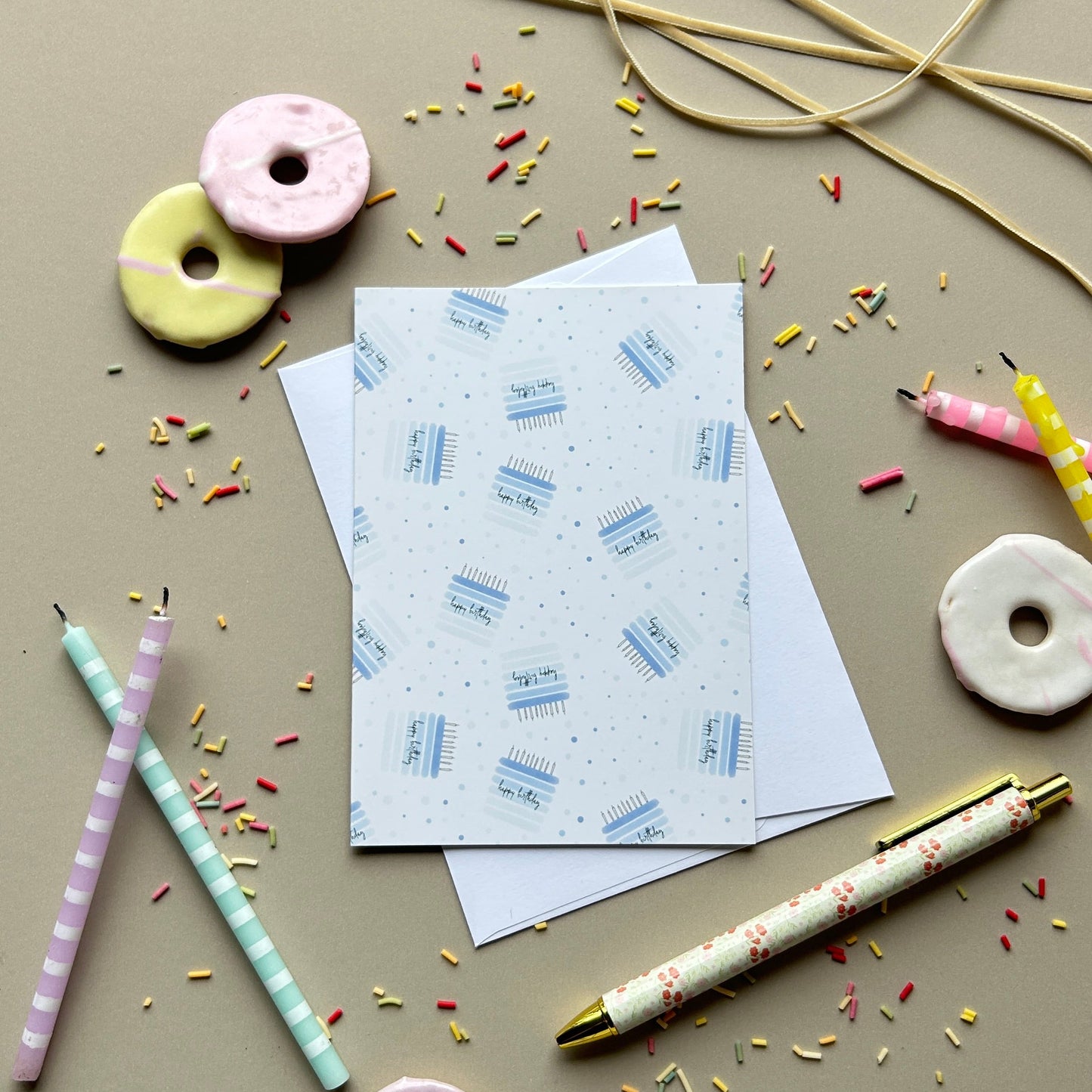 A styled flat-lay of stationery items on a warm beige background, including patterned cards, a pen, straws, and donut-shaped accessories.