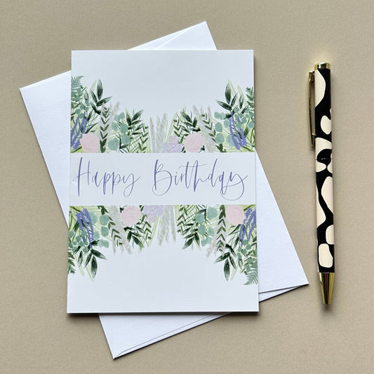 A square white birthday card with a watercolor botanical border in green, blue, and lavender, featuring the words 'Happy Birthday' in script, shown with white envelopes and a black-and-white pen on a beige background.