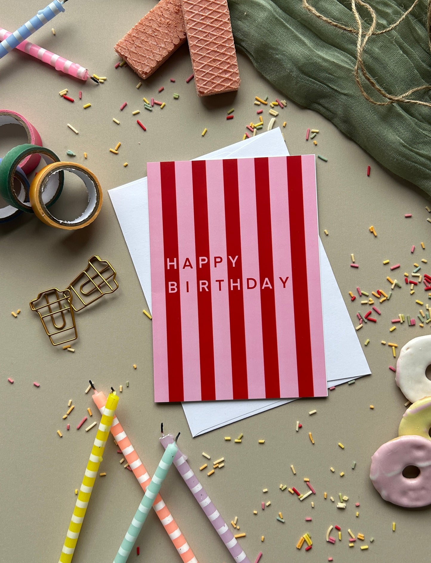 A striped birthday card with 'HAPPY BIRTHDAY' in pink text, styled in a flat lay with pastel pencils, washi tape, cookies, and confetti on a neutral background.