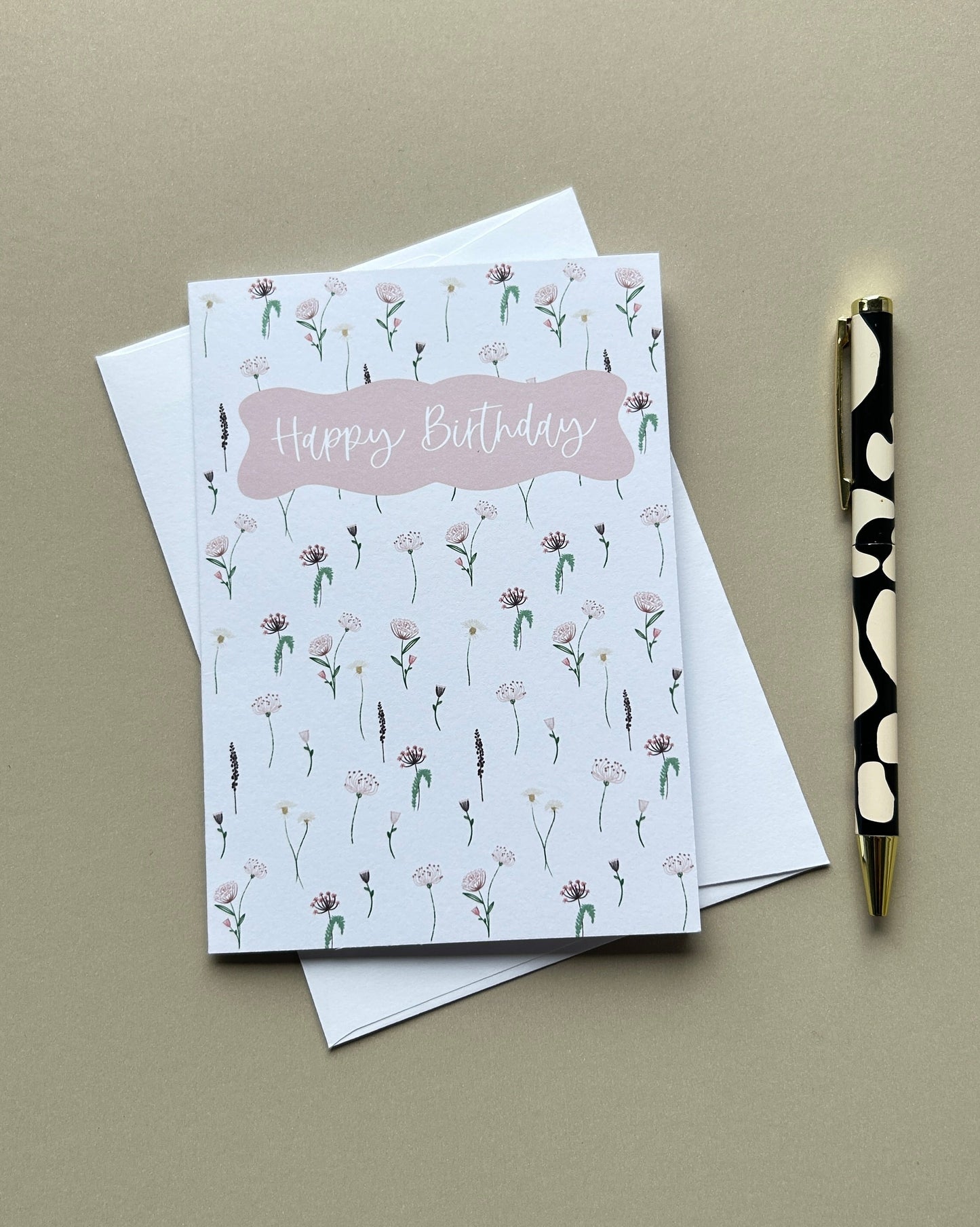 A square greeting card with a white background, a pink 'Happy Birthday' banner, and a delicate pink and green floral pattern, shown with a white envelope and a black-and-gold pen on a beige background.