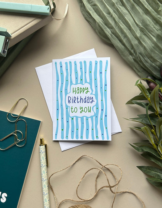 A square birthday greeting card with blue vertical stripes and the text 'Happy Birthday to You' in green and blue lettering, styled on a beige tabletop with notebooks, twine, a pen, paper clips, and greenery.
