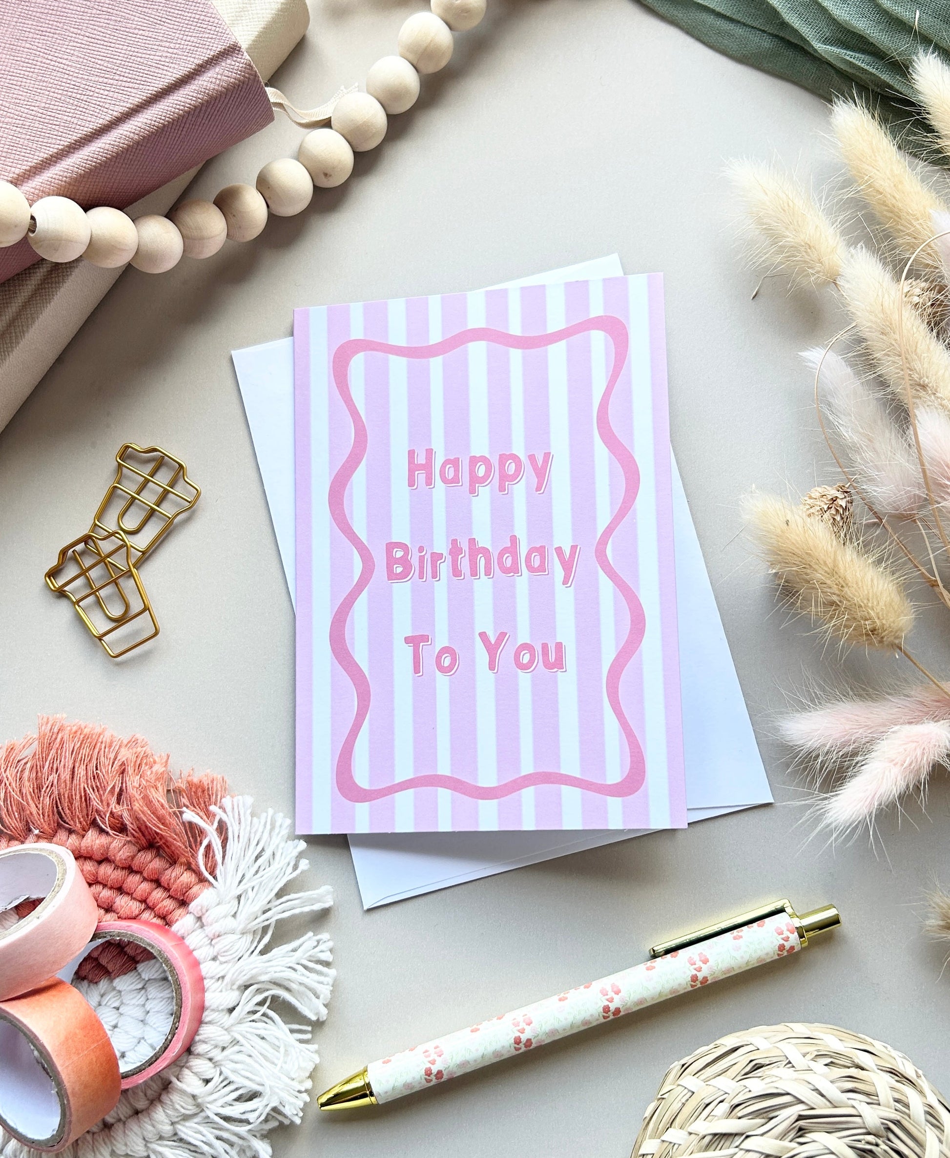 A pink and white striped birthday card with the words 'Happy Birthday To You' in pink, styled in a flat lay with wooden beads, a notebook, washi tape, a pen, and dried grass on a light gray surface.