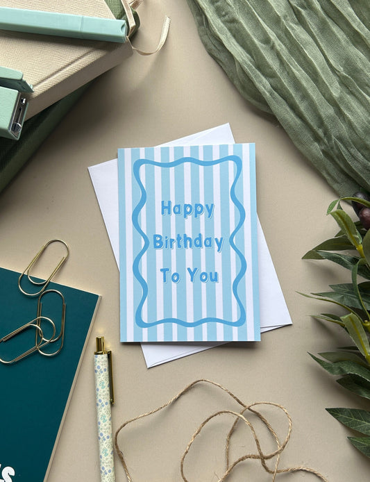 A blue and white striped 'Happy Birthday To You' greeting card displayed in a styled flat-lay arrangement with a green pen, gold paperclips, twine, and a pleated green fabric on a beige background.