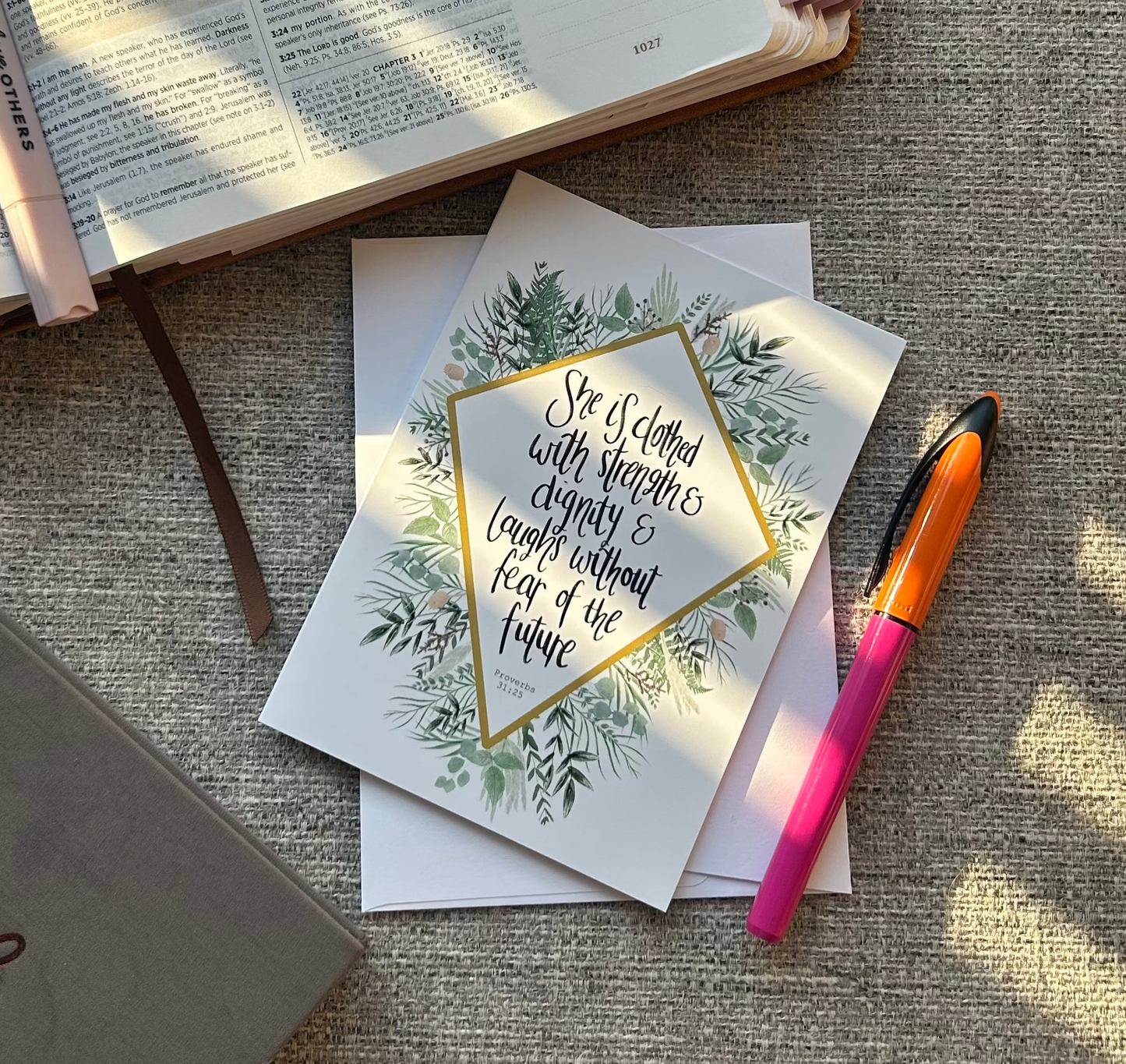 A styled flat-lay featuring a white card with a gold geometric border and green botanical illustration, placed on top of a Bible with pink tabs. A pink highlighter pen and a gray notebook are also arranged on a textured beige fabric background with natural sunlight shadows.