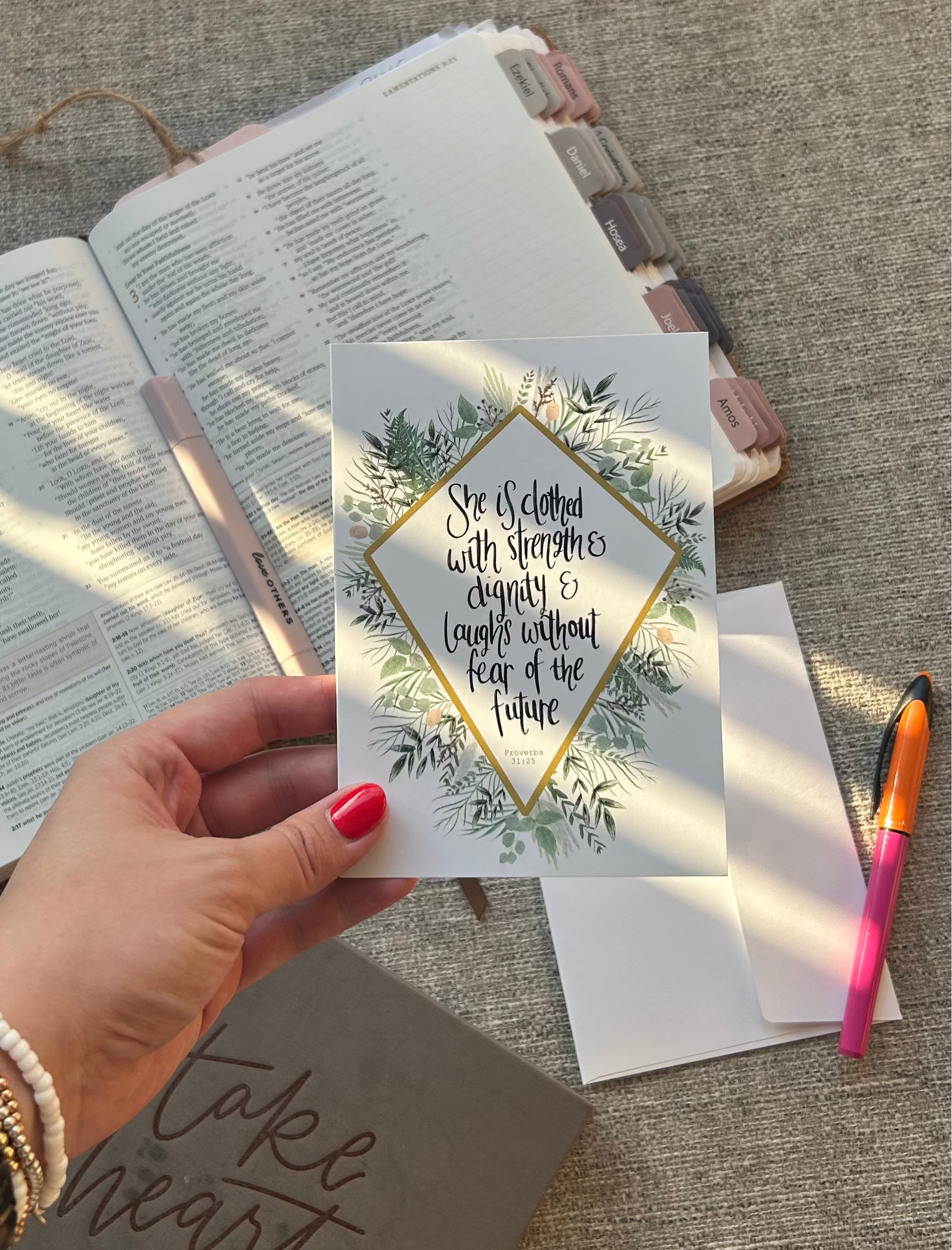 A hand with pink nail polish holds a small square card featuring a botanical border and a quote inside a diamond frame. The scene is styled on a textured gray fabric surface with an open book, a pen, and other stationery items in the background.