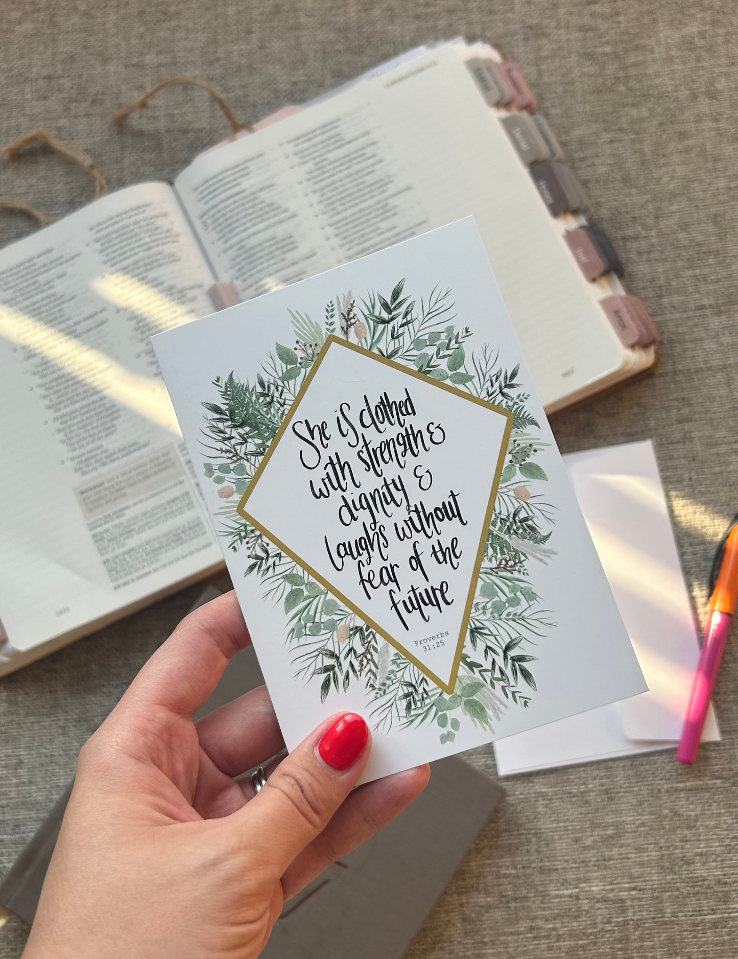 A hand with red nail polish holds a white greeting card featuring a gold diamond border, green botanical leaves, and a quote. The background shows an open book and a textured beige fabric surface.
