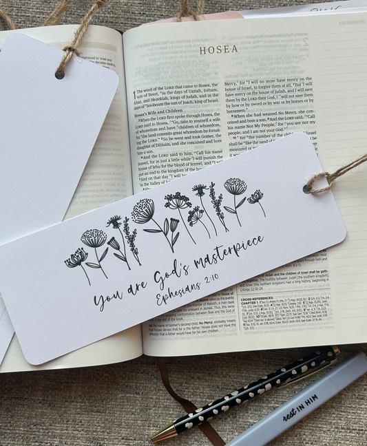 A white paper bookmark with black floral line art and the phrase 'You are God's masterpiece Ephesians 3:14' lies across an open Bible on a textured beige fabric surface. Other bookmarks, a pen, and a pencil are also visible in the styled flat-lay arrangement.