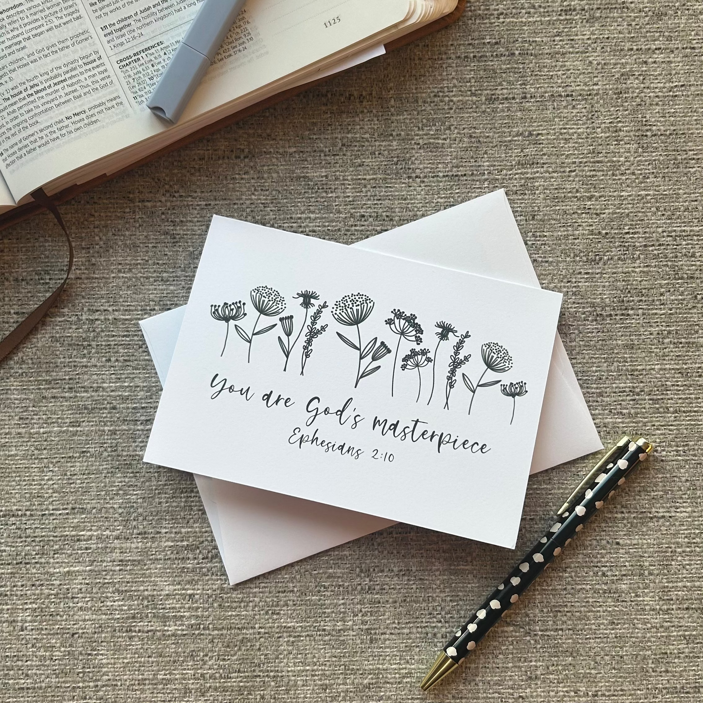 A white greeting card with black floral line art and the words 'You are God's masterpiece Ephesians 2:10' lies on top of white envelopes on a textured beige fabric surface. An open book, a gray pen, and a patterned pen are nearby as styled props.