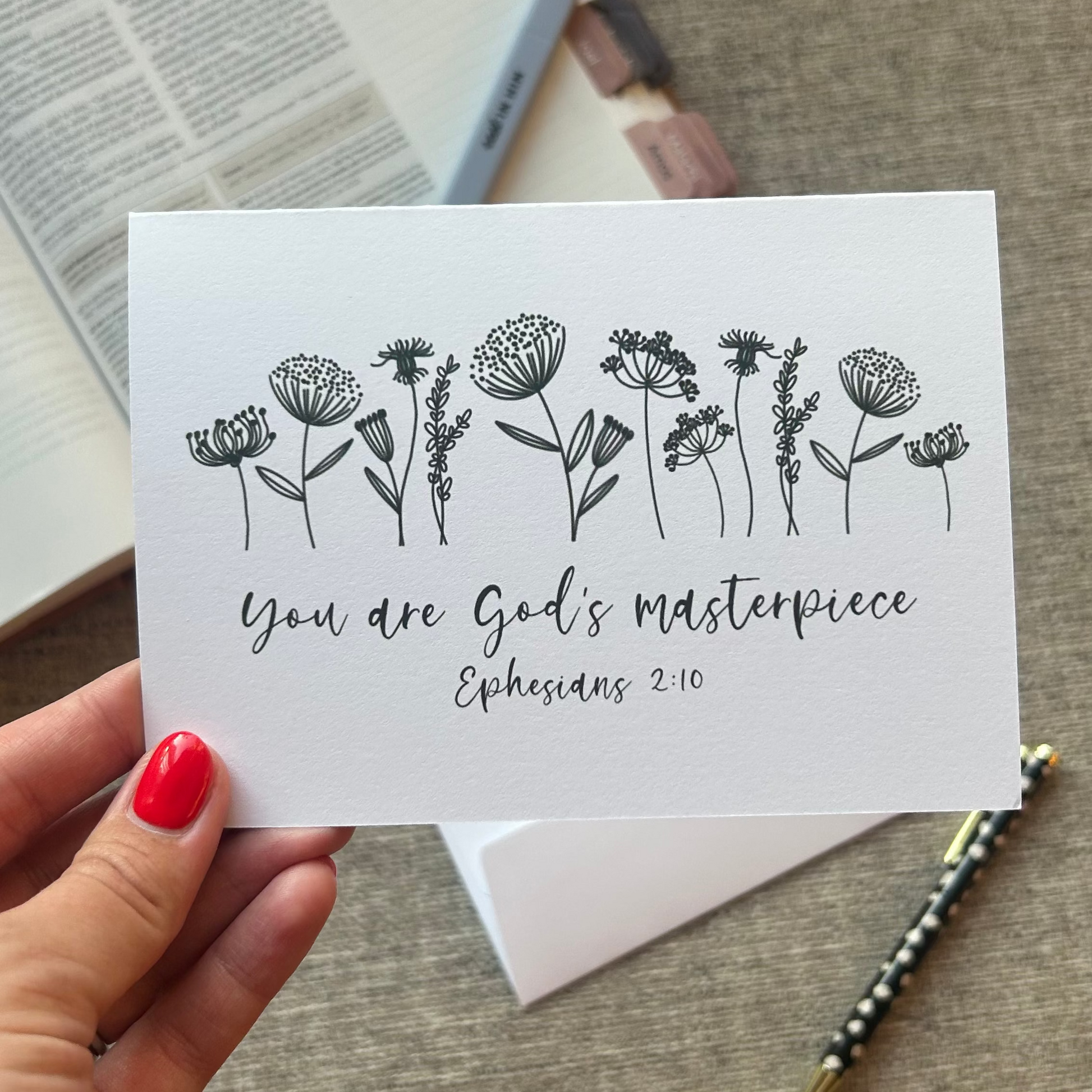 A hand with red nail polish holds a white card featuring black floral illustrations and the text 'You are God's masterpiece Ephesians 2:10'. The card is photographed on a textured gray fabric surface with an open book, envelope, and pen in the background.