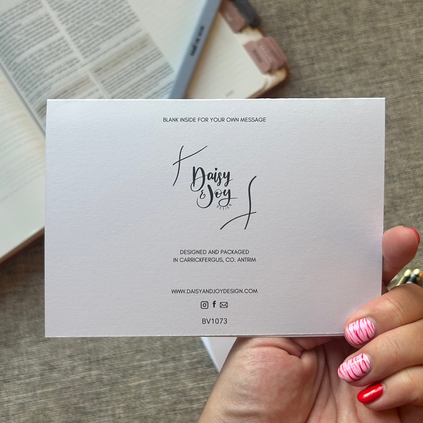 A hand holding a blank white card with elegant black script text, set against a cozy background of an open book and a textured gray fabric surface.