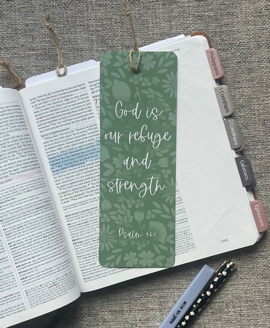 A green botanical-patterned bookmark with white script text reading 'God is our refuge and strength Psalm 34:18' resting on an open Bible, styled on a gray textured fabric background with a pen nearby.
