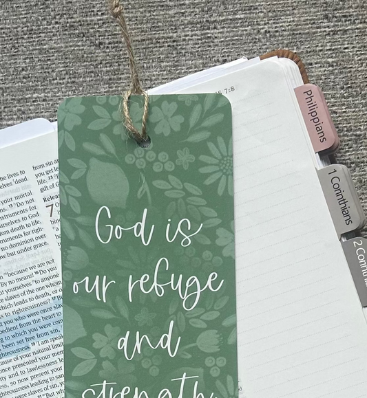 A green botanical-patterned bookmark with white script text reading 'God is our refuge and strength Psalm 46:1' resting on an open Bible, styled on a gray textured fabric background with a pen and pencil nearby.