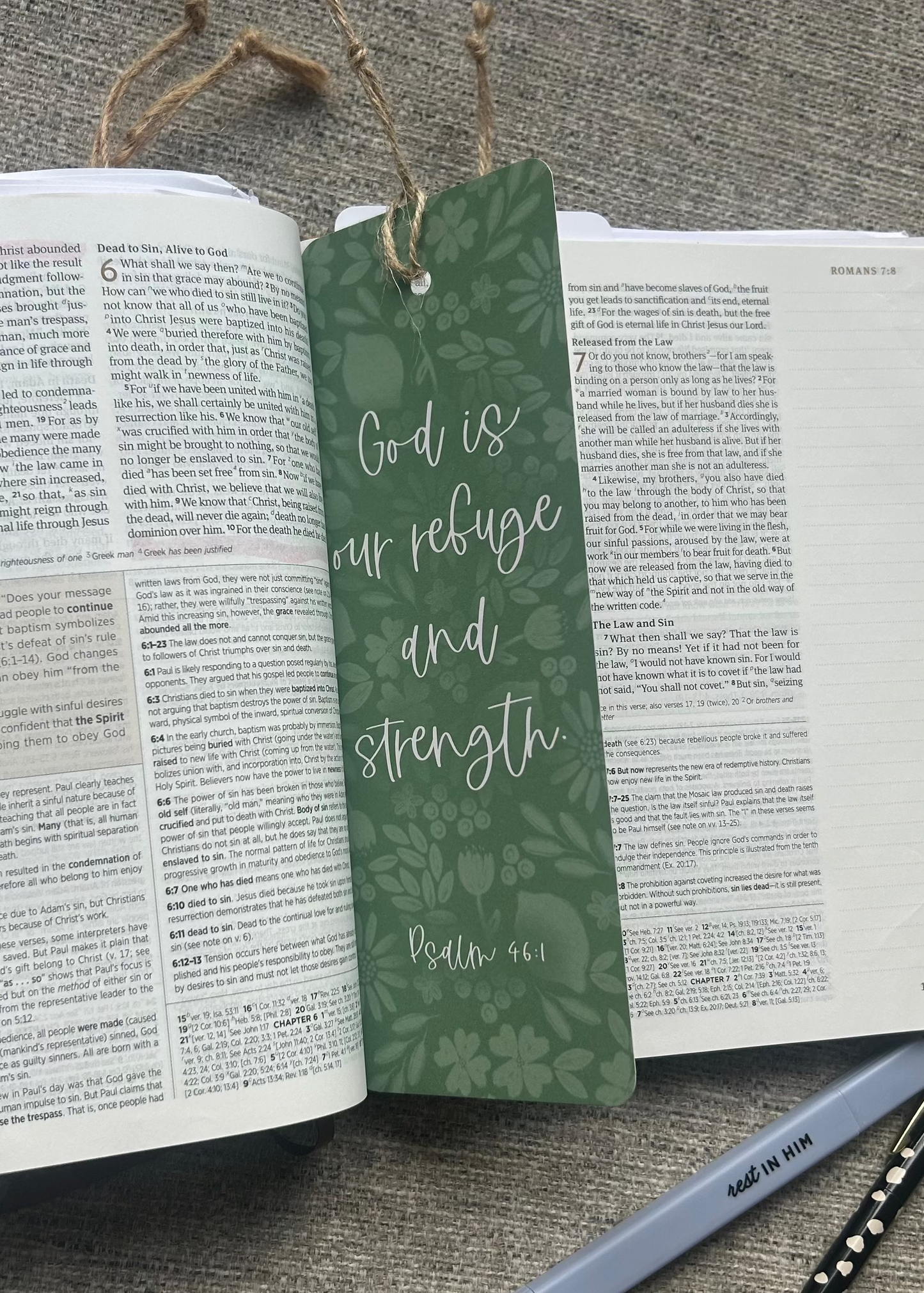 A green bookmark with a botanical pattern and the quote 'God is our refuge and strength Psalm 46:1' tied to an open Bible, styled on a gray textured fabric background with a pen and pencil.