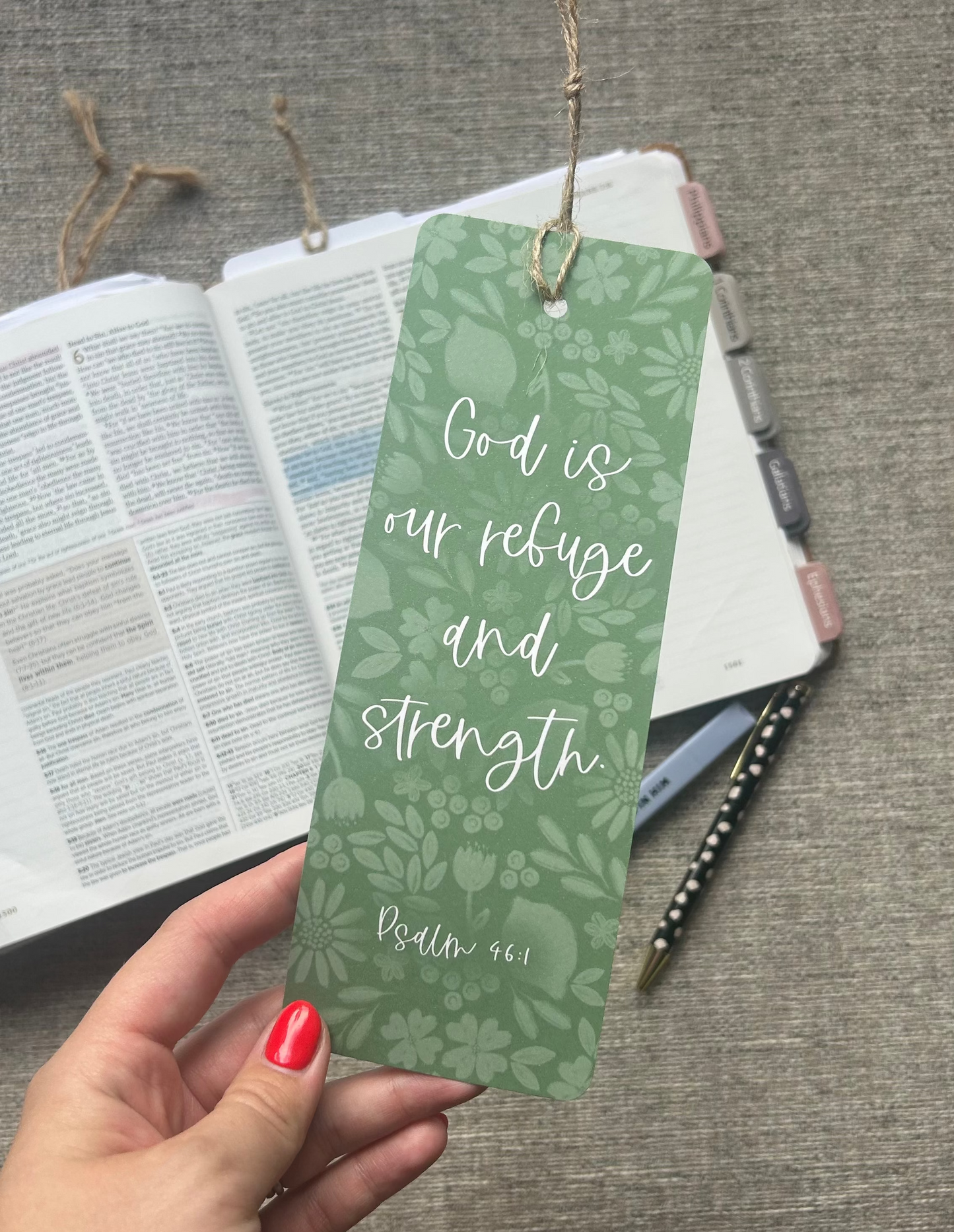 A hand with red nail polish holds a green botanical-patterned bookmark featuring the Bible verse 'God is our refuge and strength Psalm 46:1' in white script. The bookmark is shown in a styled flat-lay setting next to an open Bible, a pen, and a notebook on a textured gray fabric surface.