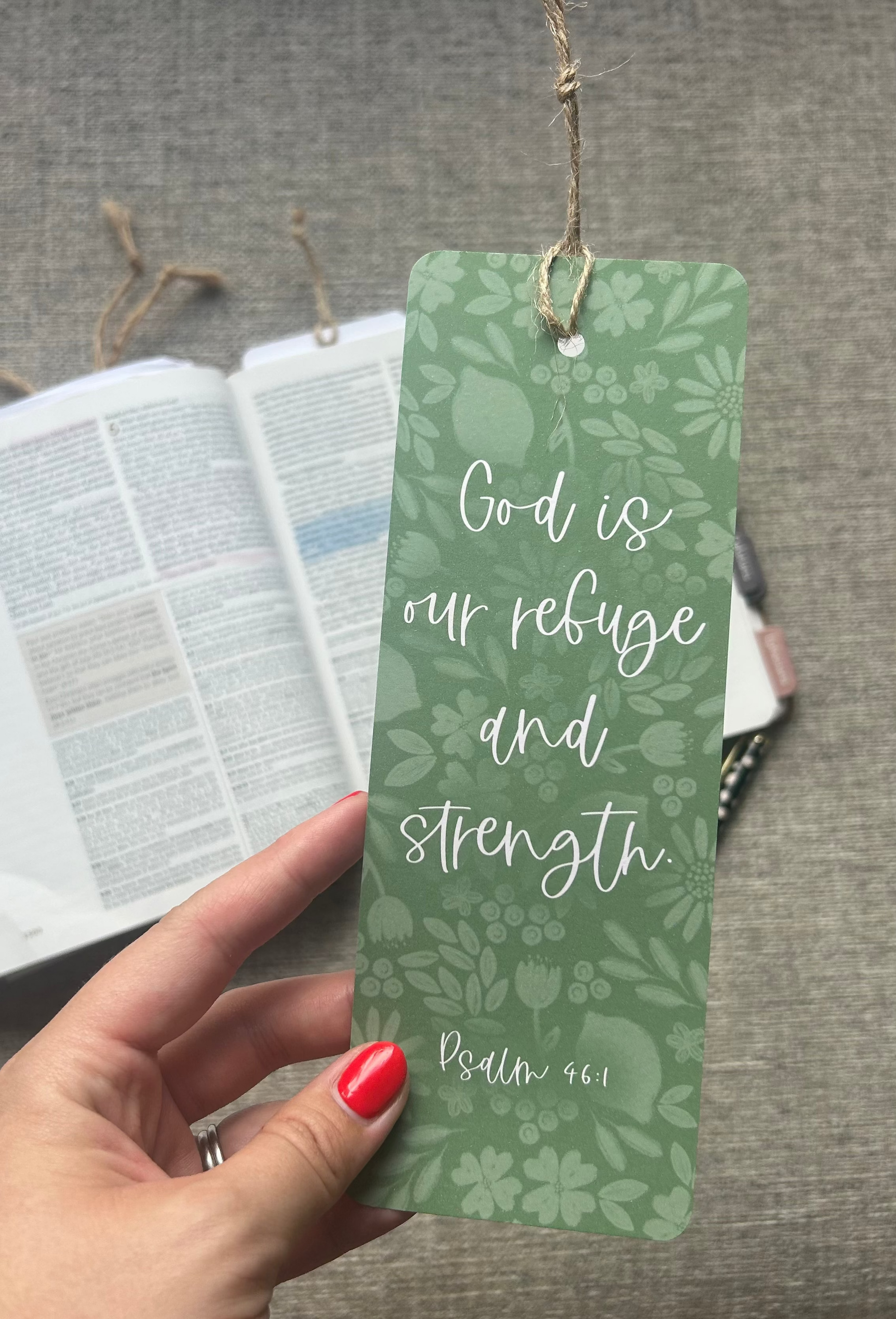 A hand with red nail polish holds a green bookmark featuring a floral pattern and the quote 'God is our refuge and strength Psalm 46:1' in white script. In the background, an open Bible lies on a gray fabric surface.