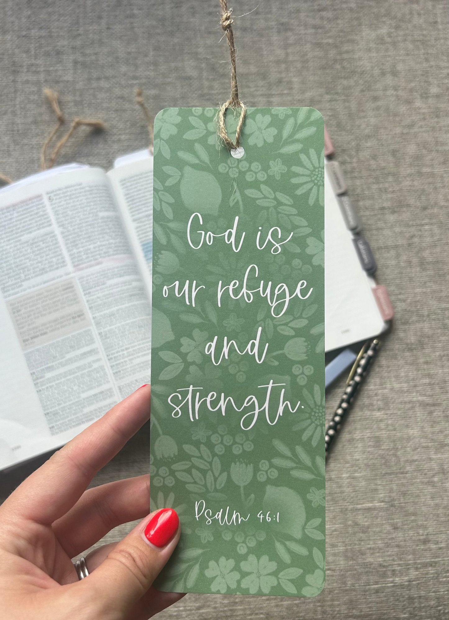 A hand with red nail polish holds a green bookmark featuring a floral pattern and the quote 'God is our refuge and strength Psalm 46:1' in white script. In the background, an open Bible and other books rest on a gray fabric surface.