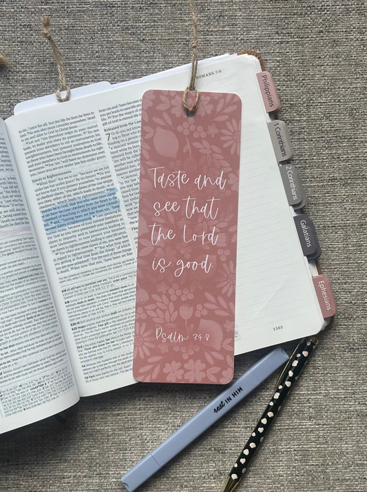 A dusty rose bookmark with a floral pattern and the Bible verse 'Taste and see that the Lord is good' from Matthew 5:3, shown resting on an open book with a pen and planner tabs nearby on a textured gray fabric background.
