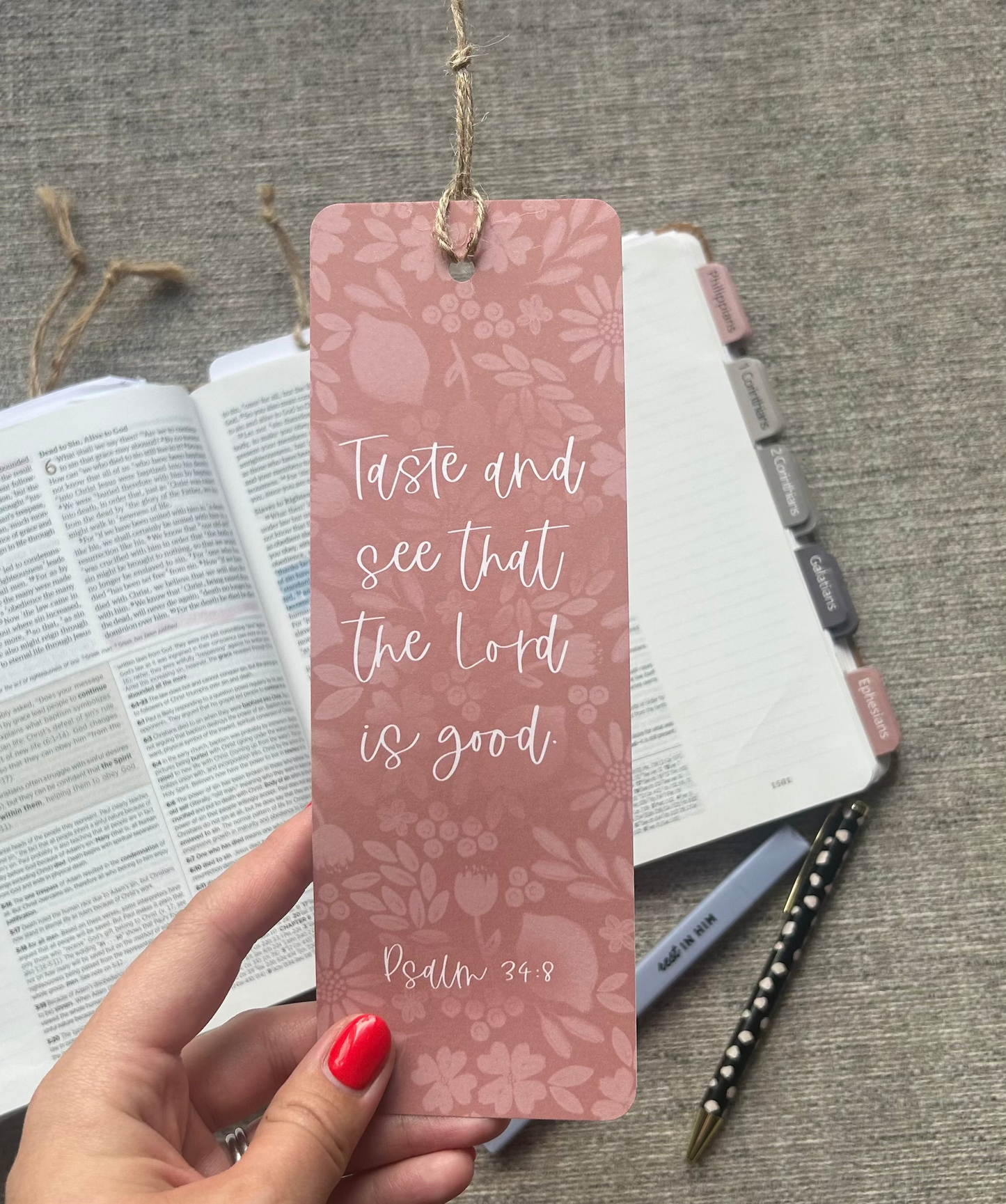 A hand with red nail polish holds a dusty pink bookmark featuring a floral pattern and the Bible verse 'Taste and see that the Lord is good' (Psalm 34:8). The bookmark is suspended by a twine string over an open book, with a planner and a pen on a gray textured fabric background.