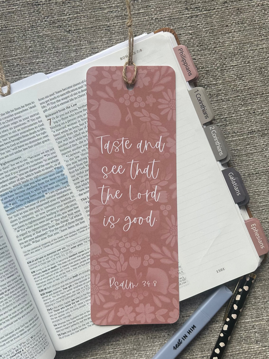 A dusty rose bookmark with a floral pattern and the quote 'Taste and see that the Lord is good Psalm 34:8' resting on an open Bible, styled with a pen and notebook on a textured gray fabric background.