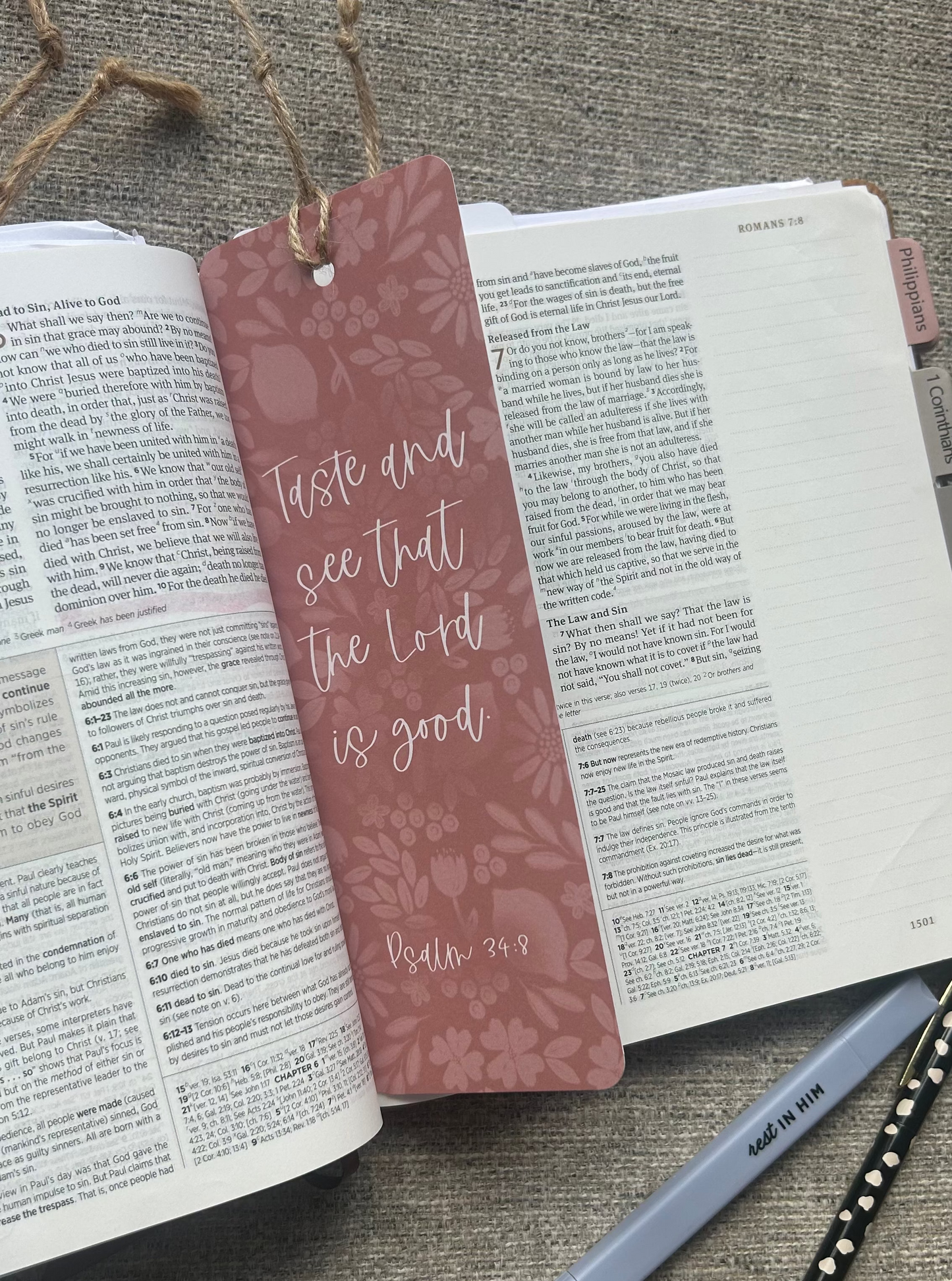 A dusty rose bookmark with a floral pattern and the Bible verse 'Taste and see that the Lord is good' (Psalm 34:8) tied to an open book on a textured gray fabric surface, with a pen nearby.